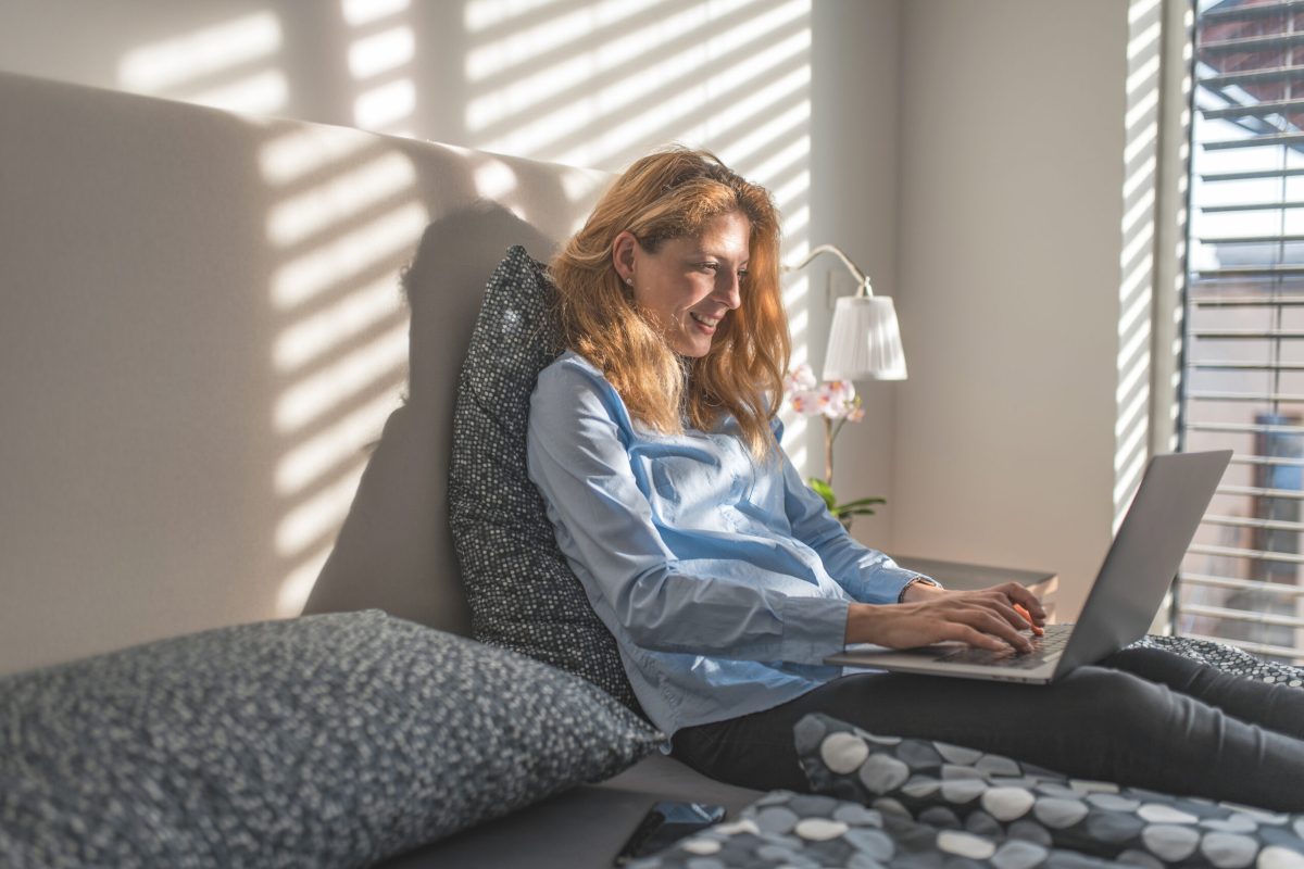 Portrait of smiling young woman using laptop in bedroom. Beautiful redhead female is sitting comfortably on bed. She is wearing casual clothing at home.